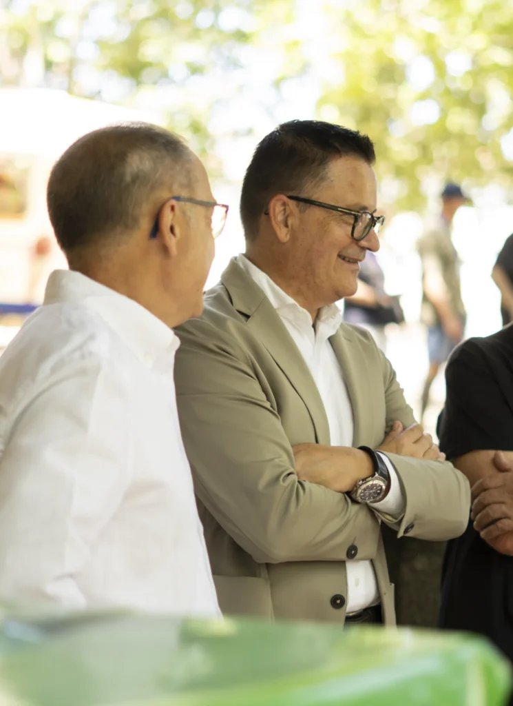 Dous homes con chaqueta e camisa branca conversando sorrindo baixo as árbores da carballeira