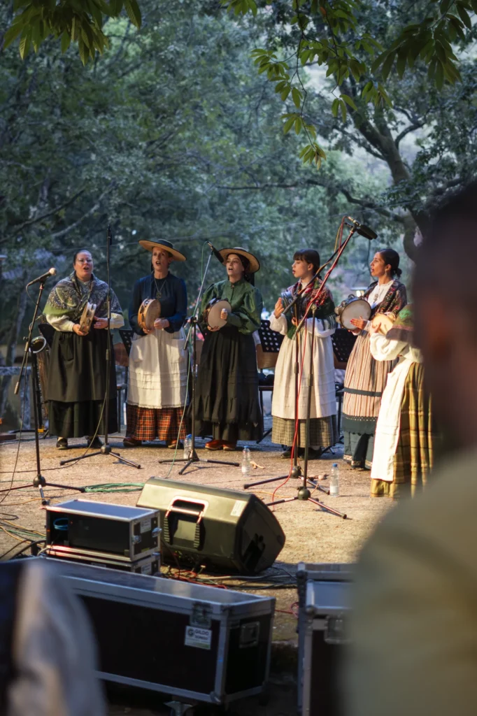 Cinco cantareiras con traxe tradicional e pandeiretas actuando entre as árbores ao solpor