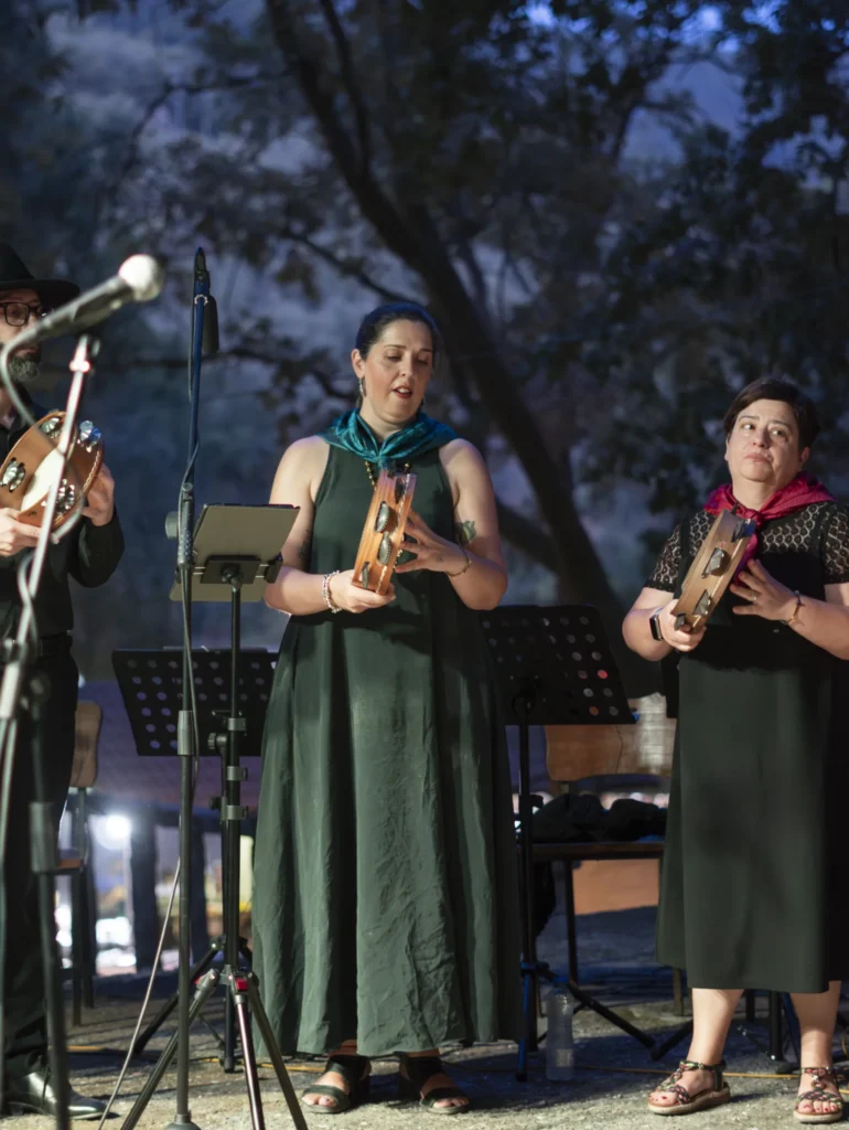 Cantareiras tocando pandeiros cadrados no escenario ao anoitecer, con atrís e micrófonos