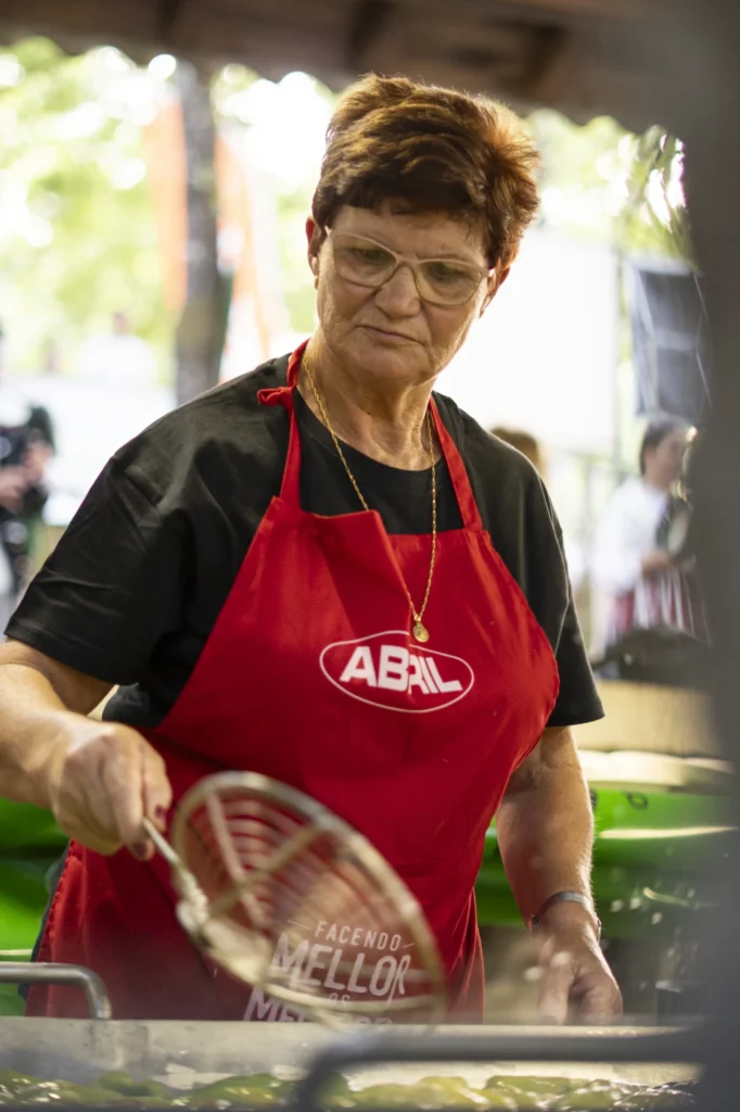 Cociñeira co mandil vermello fritindo pementos da Arnoia cunha espumadeira no posto de degustación