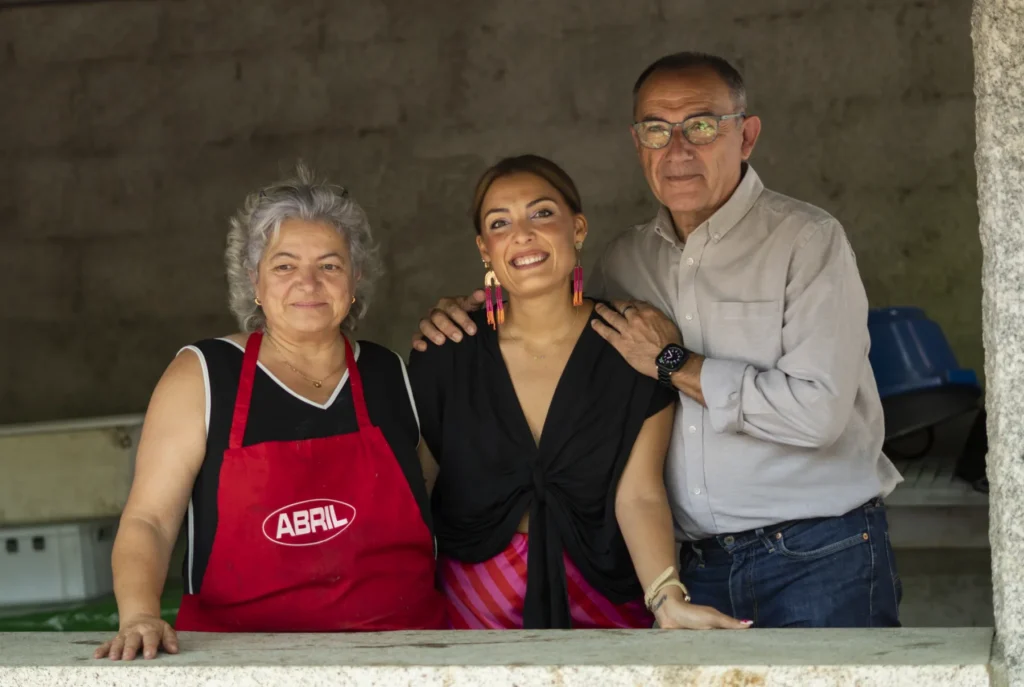 Tres persoas sorrindo no posto de degustación, unha delas co mandil vermello de cociña