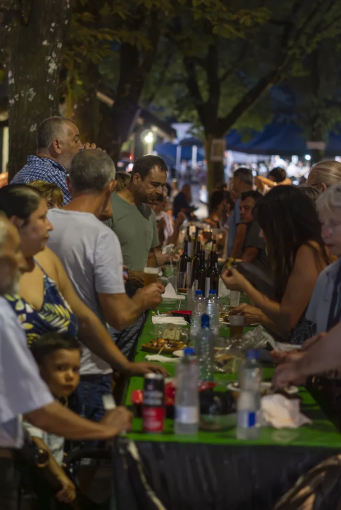 Mesa longa chea de xente ceando e compartindo viño baixo as árbores pola noite