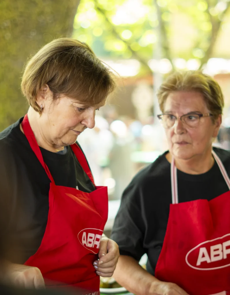 Dúas mulleres con mandil vermello atenden o posto de comida baixo as árbores do recinto