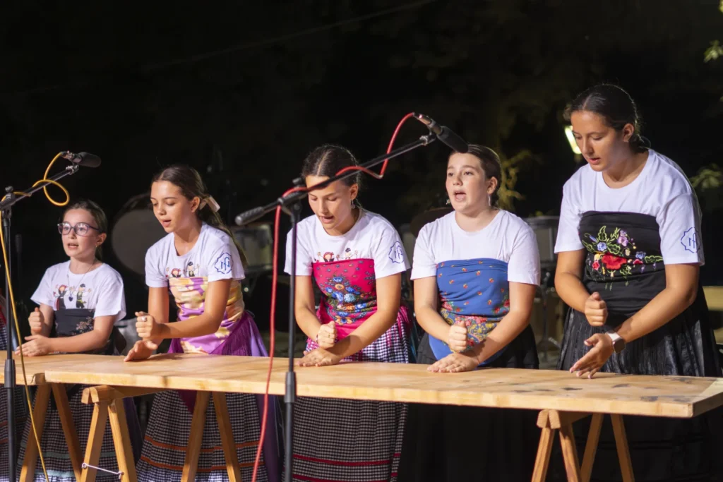 Cinco nenas con mandís tradicionais cantan e percuten sobre unha mesa de madeira no escenario