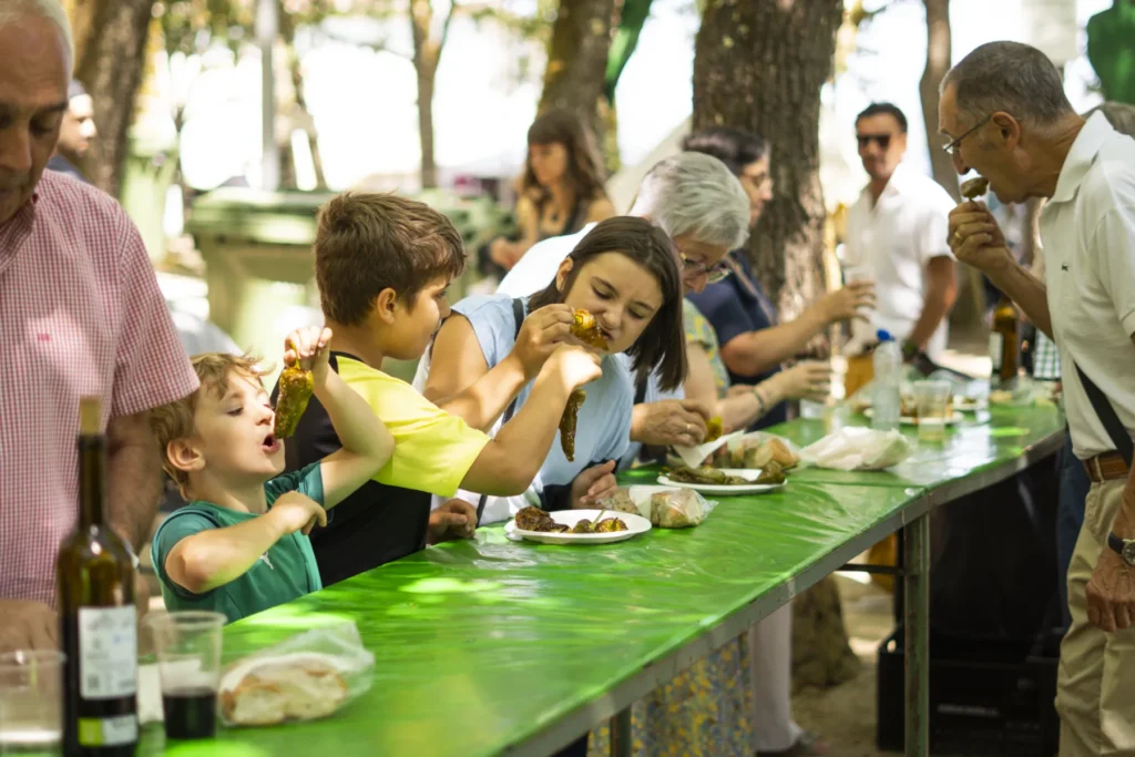 Nenos e maiores comendo pementos nunha mesa longa de mantel verde na carballeira