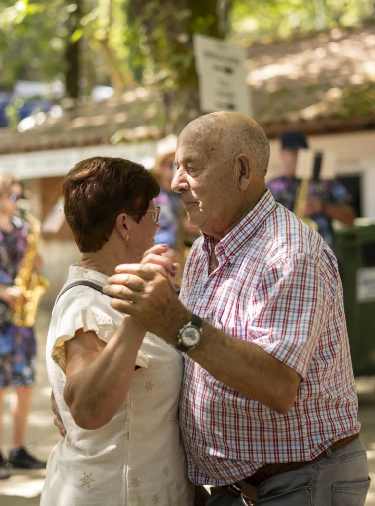 Parella maior bailando agarrada mentres un saxofonista toca ao fondo na carballeira da festa