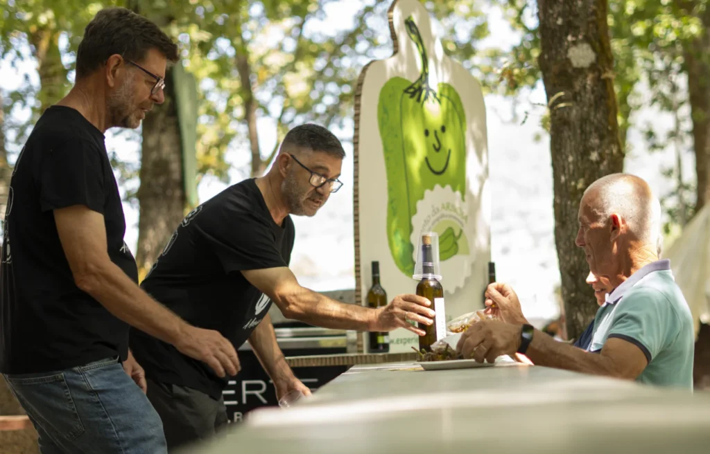Voluntario serve unha botella de viño a un visitante diante do cartel do pemento sorrinte