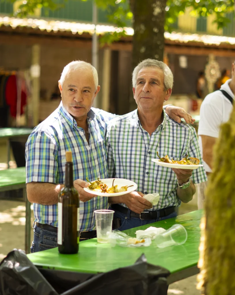 Dous veciños amosando os seus pratos de pementos xunto a unha botella de viño
