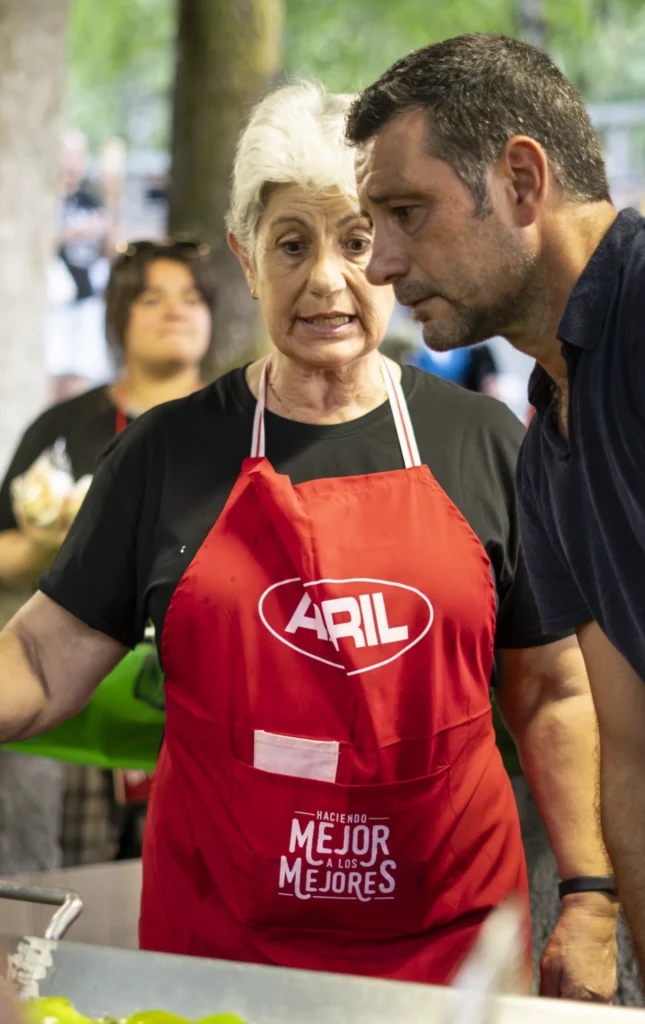 Voluntaria con mandil vermello atende o posto de pementos mentres un home lle fala