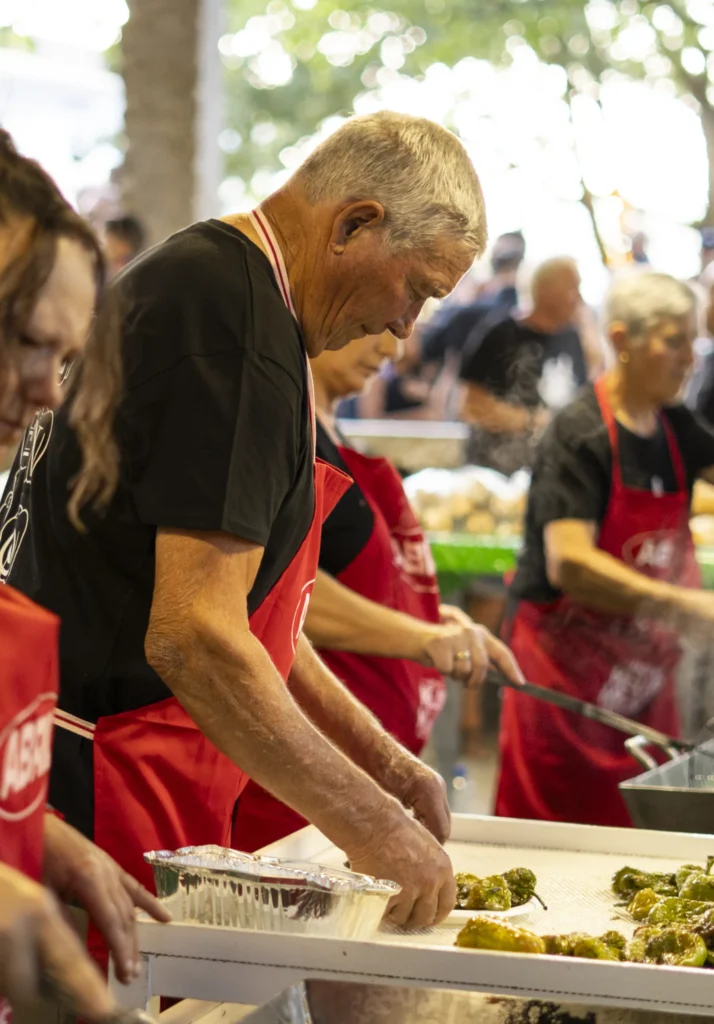 Voluntario con mandil vermello empratando pementos fritidos no posto de degustación da festa